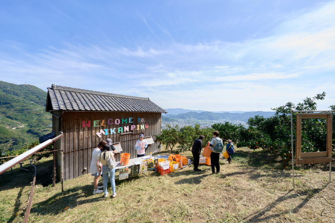 青空の下、みかん畑の丘に建つ小屋と参加者